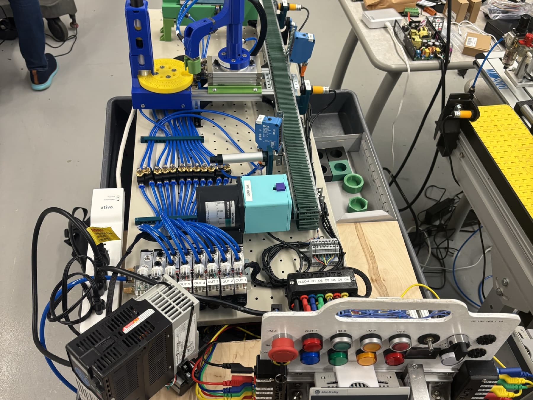 Members working on a PLC automation trainer fitted with a conveyor, pneumatics, a servo gearbox, and an operator panel
